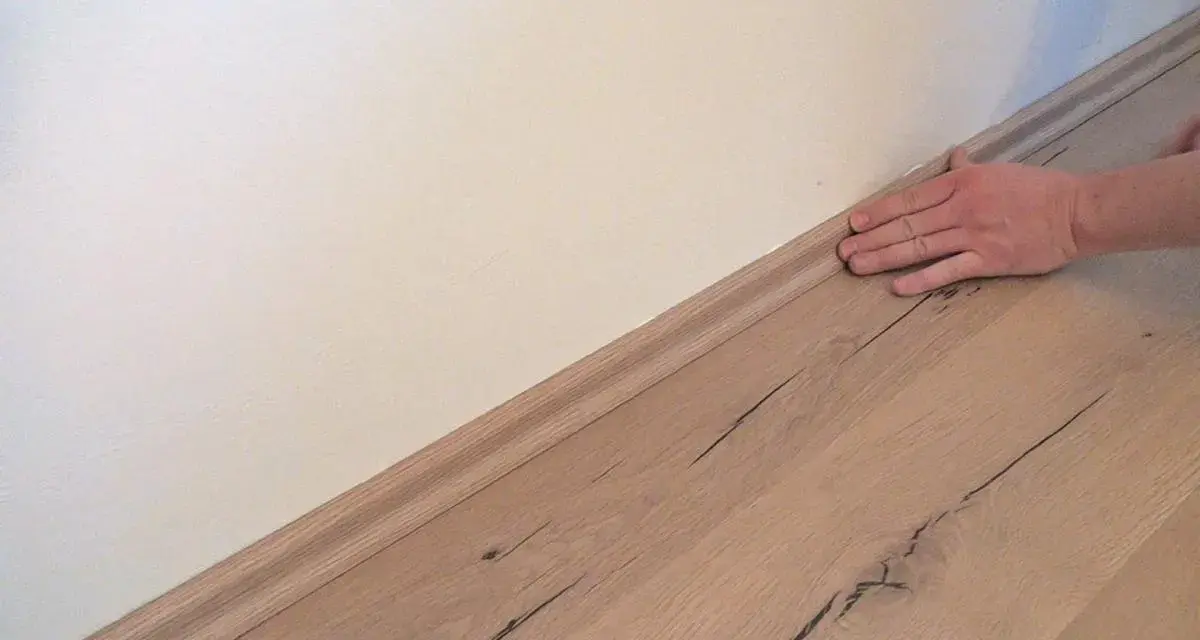 Hand drückt Sockelleisten an die Wand. Der Boden ist aus hellem Holz.