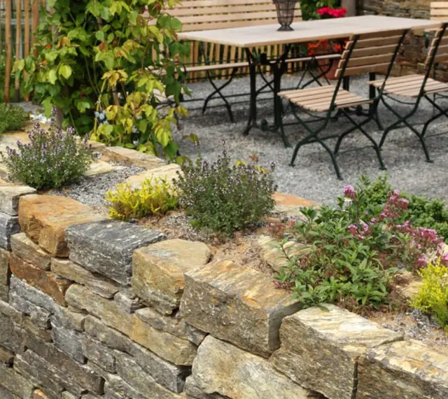 Natürliche Steinmauer mit Kräutern und Blumen, die schöne Ideen für den Garten bietet. Eine Sitzgruppe lädt zum Verweilen ein.