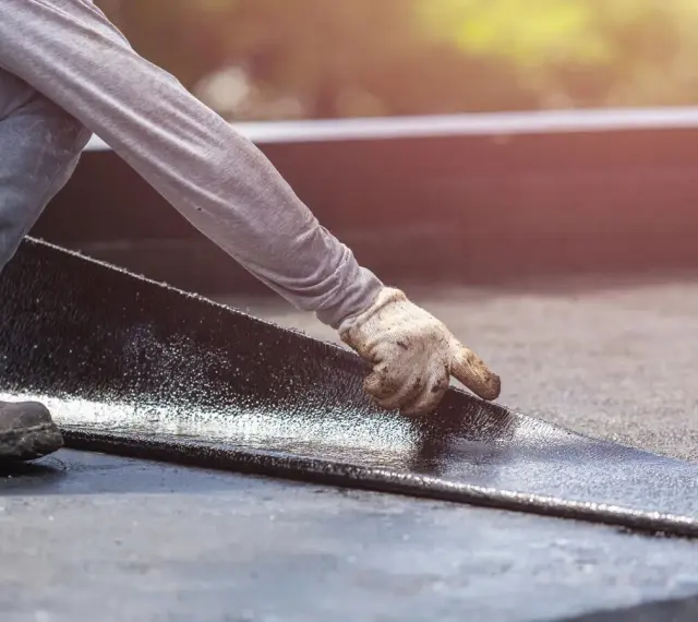 Hand mit Handschuh legt neue Dachpappe an. Alte Dachpappe entsorgen ist der nächste Schritt.