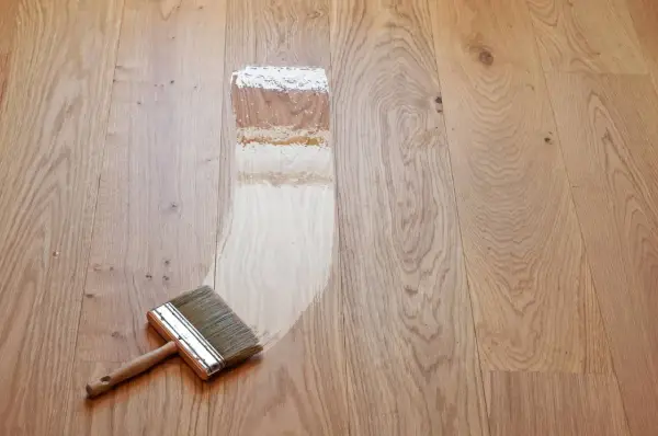 Breite Pinsel mit Holzgriff trägt Lack auf Parkett auf. Der Prozess, das Parkett zu versiegeln, zeigt den Unterschied zwischen unbehandeltem und frisch lackiertem Holz.