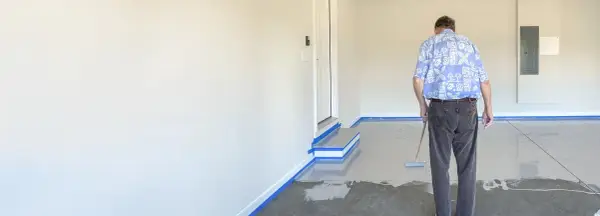 Mann beschichtet den garagenboden mit einem Roller. Der Boden ist teilweise mit einer grauen Beschichtung bedeckt, die Ränder sind mit blauer Klebefolie abgeklebt.