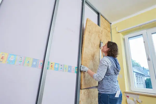 Frau baut eine Trockenbauwand, setzt Dämmmaterial in Metallständer ein.